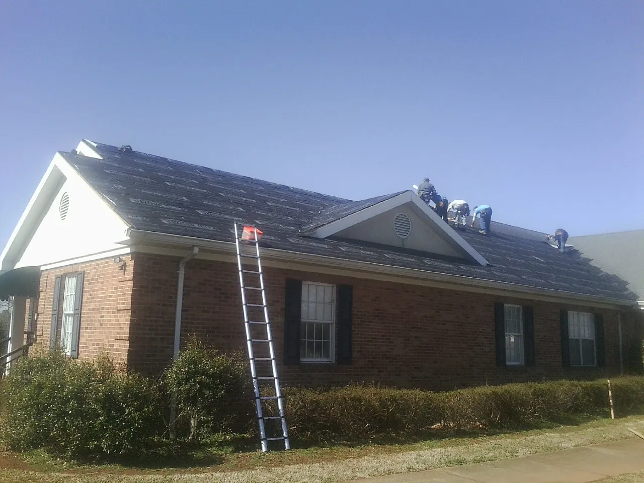 Crew tearing shingles off a brick ranch roof with a ladder set — Emergency Roof Repair work by our crew in Hales Corners