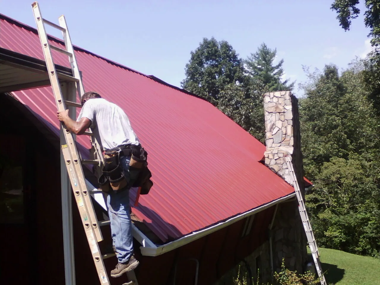 Roofer on a ladder beside a red metal roof and stone chimney — Soffit & Fascia Repair by Roofing Brothers in Hales Corners